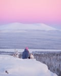 A person in a blue jacket and pink hat sits on a snowy hilltop with a small white dog, overlooking a vast snowy landscape and distant mountains under a pink and purple sky.