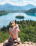 Woman with a dog sits on a rock, overlooking a lake with an island and mountains in the background.
