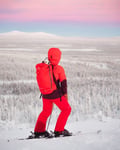 Person in red winter gear with skis stands on snowy hill, overlooking a snowy forest at sunset.
