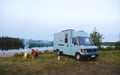 A person tends a campfire near a lake, with a light blue camper van parked on grass nearby. A small dog sits in front of the van, and trees line the lake in the background under a cloudy sky.