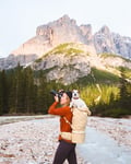A woman stands by a rocky stream with tall pine trees and mountains in the background, holding a camera to her eye. She carries a small white dog in a beige backpack.